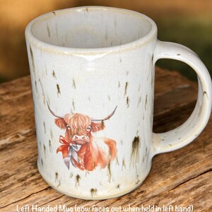 May include: Cream-colored ceramic mug with a watercolor illustration of a Highland cow wearing a red bow. The mug has a speckled finish and a curved handle. Text on the mug reads: Left Handed Mug (cow faces out when held in left hand).
