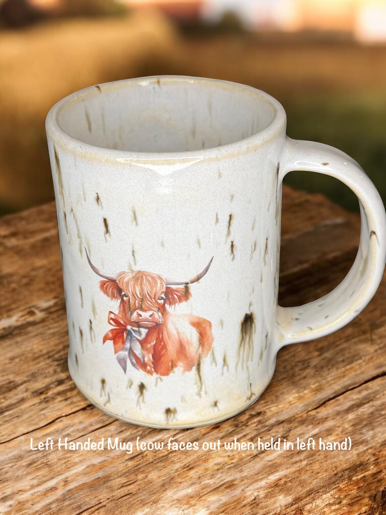 May include: Cream-colored ceramic mug with a watercolor illustration of a Highland cow wearing a red bow. The mug has a speckled finish and a curved handle. Text on the mug reads: Left Handed Mug (cow faces out when held in left hand).