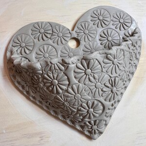May include: Gray heart-shaped wall pocket with a floral daisy pattern. The ceramic pocket has a small hole at the top for hanging. The front of the pocket is decorated with embossed daisies.