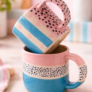 Puede incluir: Dos tazas de cerámica apiladas, con una combinación de colores rosa, azul y blanco. Las tazas tienen un diseño a rayas con lunares negros y asas redondeadas. Las tazas son adecuadas para café o té.