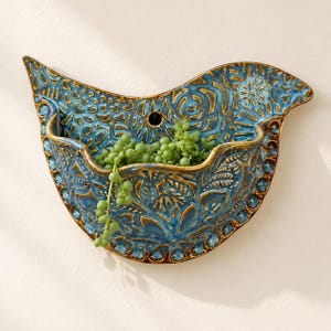 May include: A bird-shaped wall planter made of glazed ceramic with a blue and bronze finish. The surface features intricate embossed floral and leaf patterns. A small green succulent plant is tucked inside the curved pocket.
