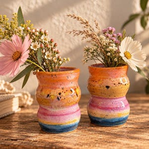 May include: Two ceramic vases with a wavy, hourglass-like silhouette, featuring a speckled glaze in horizontal bands of orange, pink, yellow, and blue. Each vase holds a bouquet of assorted wildflowers, including pink and white cosmos flowers.