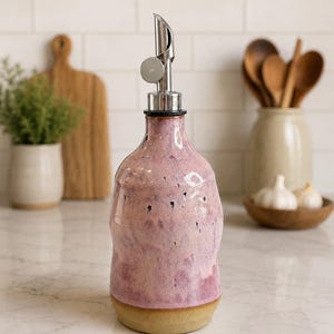 Puede incluir: Un dispensador de aceite de cer&aacute;mica con esmalte moteado rosa y morado, base beige sin esmaltar y vertedor de acero inoxidable. La palabra Bougainvillea est&aacute; impresa en blanco en la parte inferior de la botella de cuerpo redondeado.