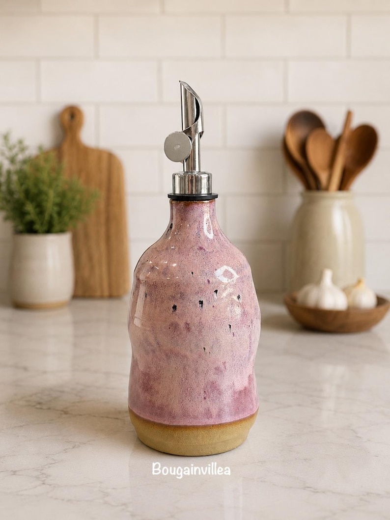 Puede incluir: Un dispensador de aceite de cer&aacute;mica con esmalte moteado rosa y morado, base beige sin esmaltar y vertedor de acero inoxidable. La palabra Bougainvillea est&aacute; impresa en blanco en la parte inferior de la botella de cuerpo redondeado.