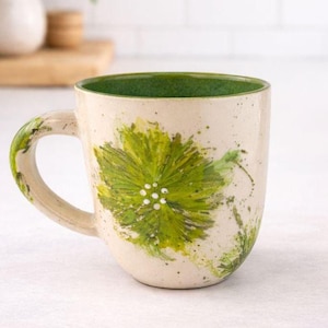 Könnte beinhalten: Cremefarbene Keramiktasse mit grünem Innenraum und Henkel. Die Tasse hat ein großes, handgemaltes grünes Blumendesign mit weißen Akzenten. Grüne Farbspritzer ergänzen das Design.