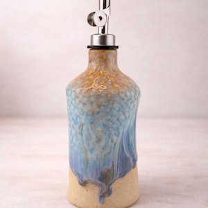 Puede incluir: Una botella dispensadora de aceite de cer&aacute;mica con un esmalte texturizado y moteado en tonos azul, bronceado y crema. La botella presenta un cuerpo cil&iacute;ndrico con cuello c&oacute;nico y un vertedor de acero inoxidable con tapa met&aacute;lica abatible.