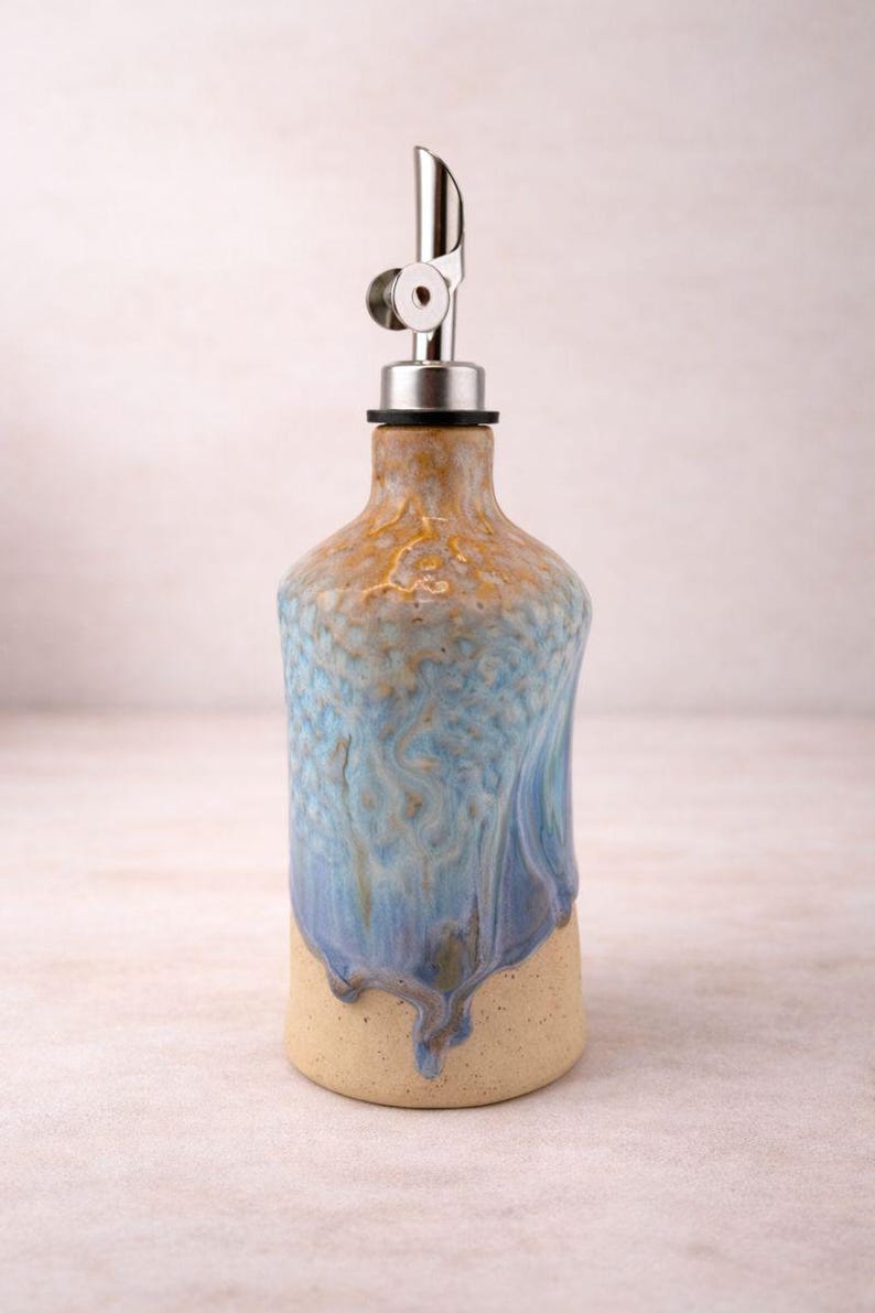 Puede incluir: Una botella dispensadora de aceite de cer&aacute;mica con un esmalte texturizado y moteado en tonos azul, bronceado y crema. La botella presenta un cuerpo cil&iacute;ndrico con cuello c&oacute;nico y un vertedor de acero inoxidable con tapa met&aacute;lica abatible.