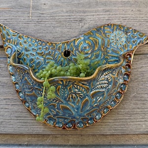 Peut inclure: Une jardinière murale en céramique en forme d'oiseau avec une finition émaillée bleue et brune, ornée de motifs floraux et géométriques en relief. L'objet présente une ouverture centrale pour les plantes et un trou pour la fixation.