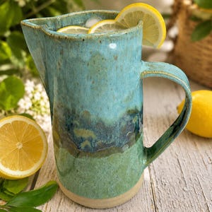 Puede incluir: Una jarra de cerámica hecha a mano con un esmalte reactivo en tonos turquesa, azul oscuro y verde. El recipiente tiene un pico vertedor ancho, un asa curva y una base de color claro. Está llena de hielo y decorada con rodajas de limón.