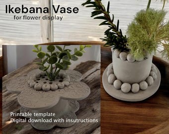 May include: A grey ceramic flower vase shaped like a flower with a decorative heart pattern. The vase has multiple holes for flower stems. The text "Ikebana Vase for flower display" is printed above the vase. The vase is on a wooden surface.