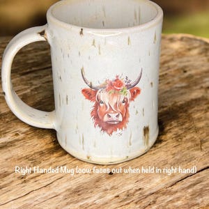 May include: Cream-colored ceramic mug featuring a watercolor illustration of a Highland cow with a floral crown. The mug has a large handle and the text "Right Handed Mug (cow faces out when held in right hand)" is printed on the mug.