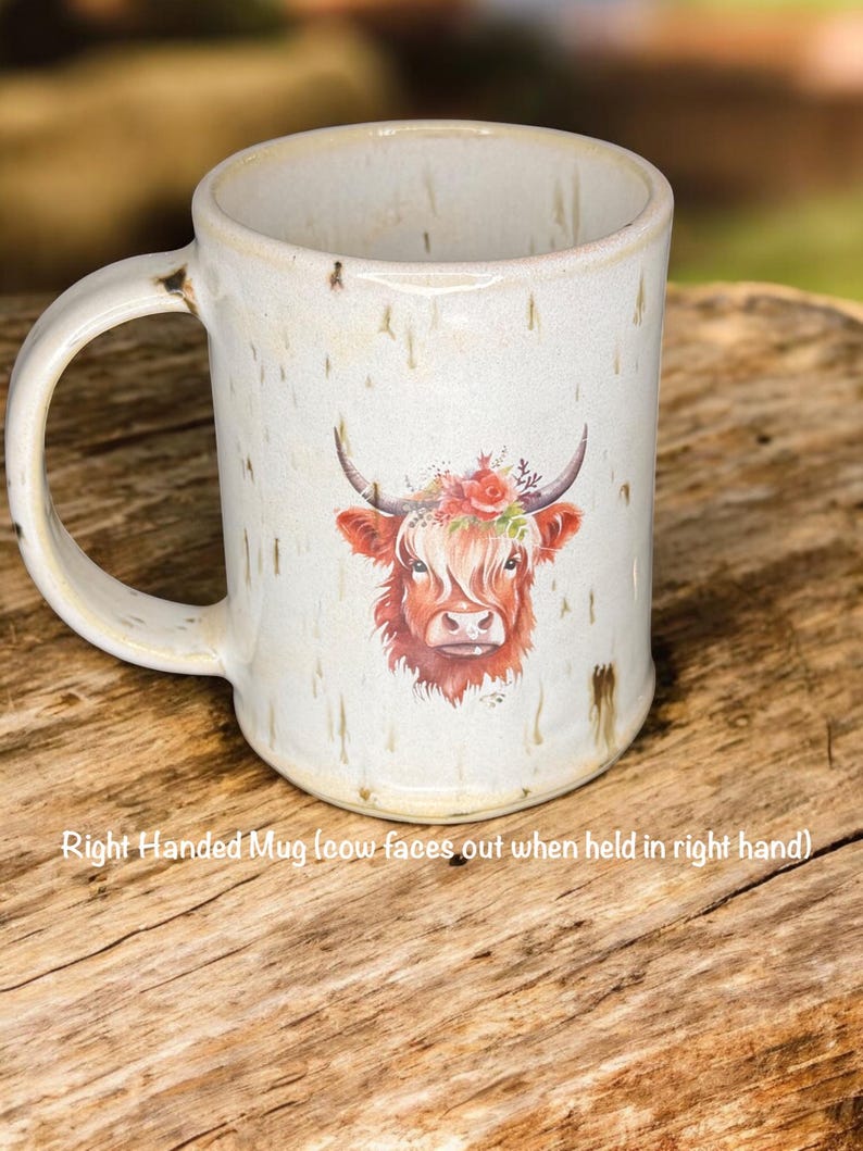May include: Cream-colored ceramic mug featuring a watercolor illustration of a Highland cow with a floral crown. The mug has a large handle and the text "Right Handed Mug (cow faces out when held in right hand)" is printed on the mug.
