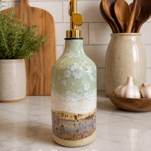 Puede incluir: Un dispensador de aceite de cer&aacute;mica con un vertedor de metal dorado, que presenta un esmalte de varios tonos que transita de verde salvia en la parte superior a blanco, marr&oacute;n y azul en la base. El texto "Sage Bloom - special edition" est&aacute; impreso abajo.