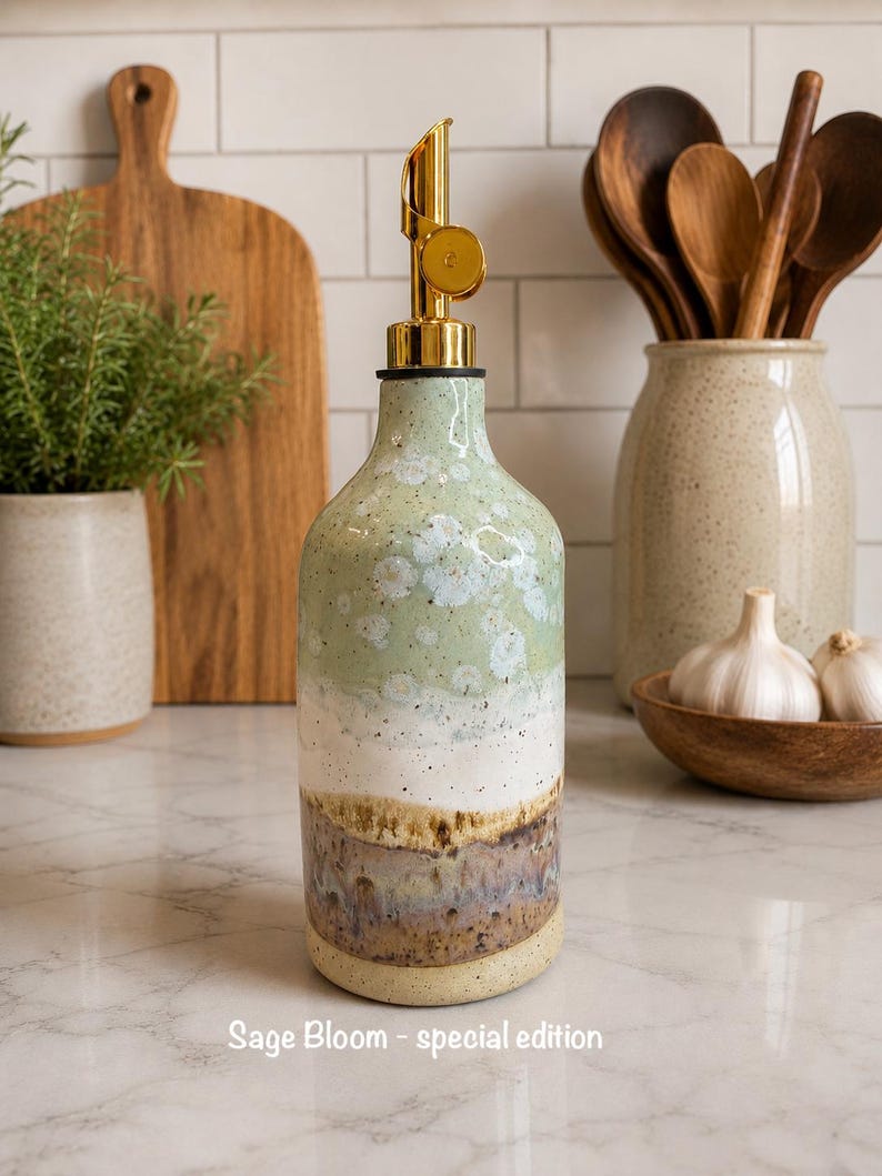 Puede incluir: Un dispensador de aceite de cer&aacute;mica con un vertedor de metal dorado, que presenta un esmalte de varios tonos que transita de verde salvia en la parte superior a blanco, marr&oacute;n y azul en la base. El texto "Sage Bloom - special edition" est&aacute; impreso abajo.