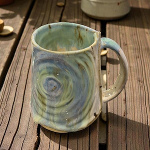 May include: A handcrafted ceramic mug with a distinctive swirl design in blue, green, and tan hues. The mug features a large handle and an irregular form, indicating it is a piece of pottery.