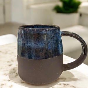 May include: A handcrafted ceramic mug. The upper half has a dark blue, textured glaze, while the base is matte brown. The mug features a rounded handle and is suitable for hot drinks.