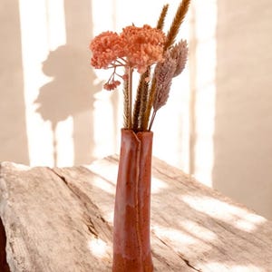 Könnte beinhalten: Eine terracottafarbene Vase mit strukturierter Oberfläche hält einen Strauß getrockneter Blumen, darunter rosa und beige Blüten und lange, dünne Gräser.
