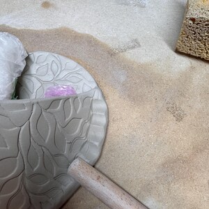 May include: A partially finished ceramic bowl with a leaf pattern, a wooden rolling pin, and a sponge. The bowl is gray and has a detailed leaf design. The rolling pin is light brown. The sponge is yellow.
