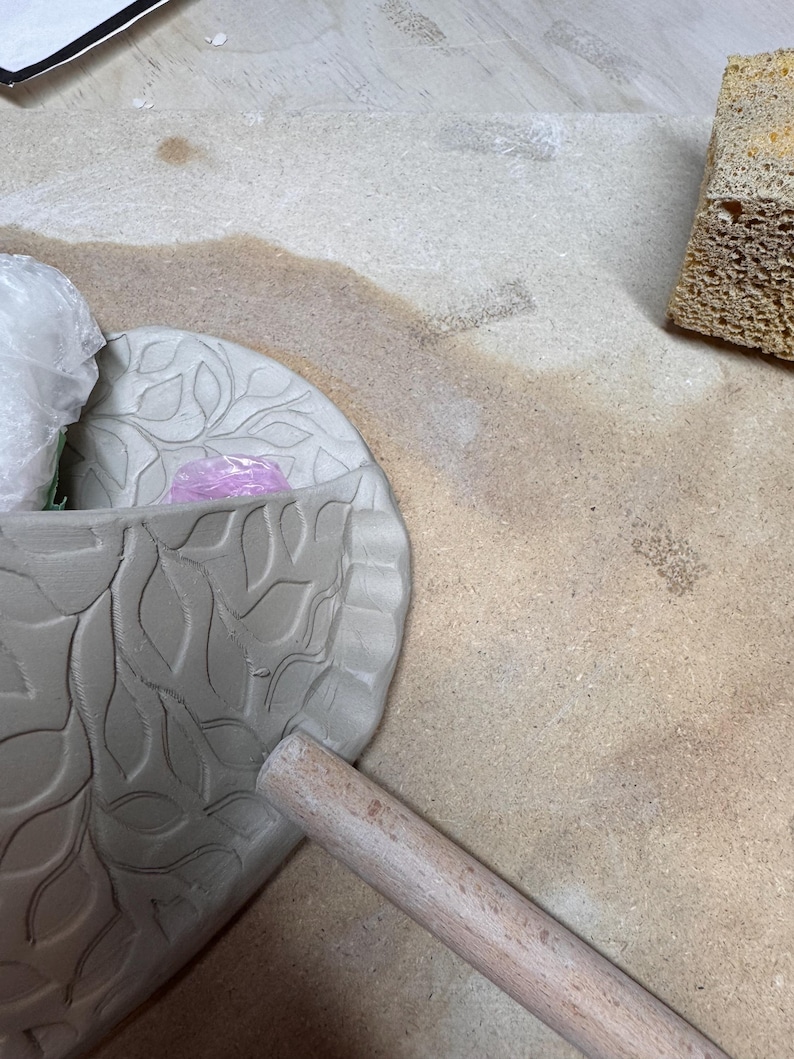 May include: A partially finished ceramic bowl with a leaf pattern, a wooden rolling pin, and a sponge. The bowl is gray and has a detailed leaf design. The rolling pin is light brown. The sponge is yellow.