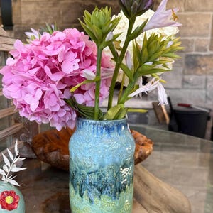 Op de afbeelding: Een keramische vaas met een getextureerde, geglazuurde afwerking in tinten blauw, groen en wit bevat een boeket roze hortensia's en andere bloemen. De vaas heeft een cilindrische vorm en staat op een glazen tafel.