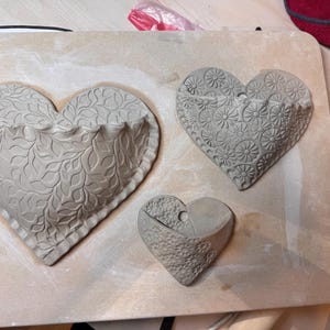 May include: Three gray, heart-shaped wall planters with textured designs. The largest heart features a leaf pattern, the middle one has a floral design, and the smallest has a textured surface. They are arranged on a light-colored surface.