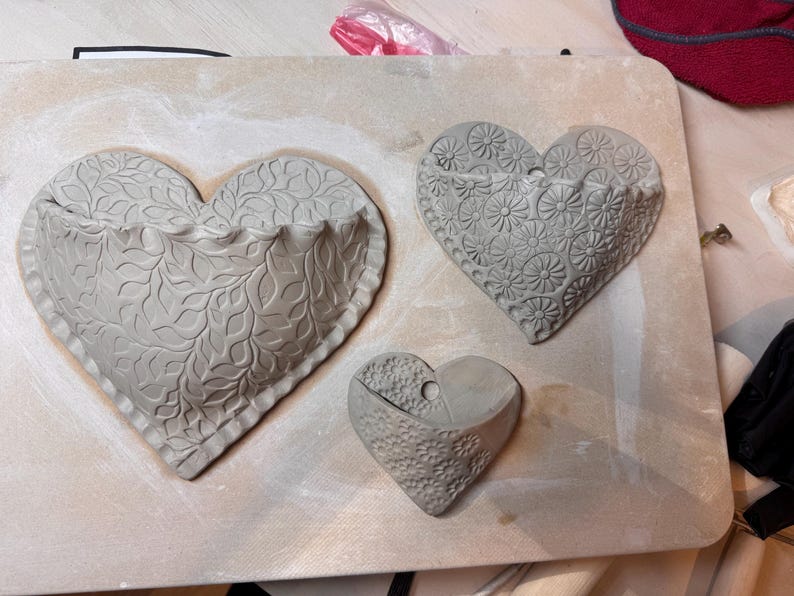 May include: Three gray, heart-shaped wall planters with textured designs. The largest heart features a leaf pattern, the middle one has a floral design, and the smallest has a textured surface. They are arranged on a light-colored surface.