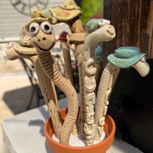 May include: A terracotta pot filled with whimsical ceramic garden stakes. The stakes are shaped like smiling snakes wearing hats, with various colours and designs. The stakes are a unique and decorative addition to any garden.
