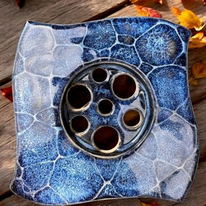 Op de afbeelding: Een vierkant decoratief keramisch stuk met een blauw en wit gevlekt patroon. Het midden heeft een cirkelvormig metalen element met meerdere gaten. Het stuk ligt op een houten oppervlak, met de tekst "BBSQ-1" zichtbaar.