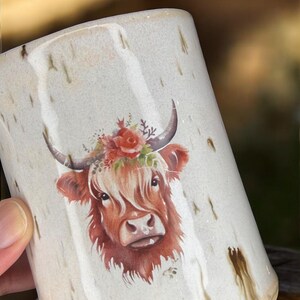 May include: Cream-colored ceramic mug featuring a Highland cow illustration. The cow has reddish-brown fur, a floral crown, and curved horns. The mug has a speckled design with brown drips.