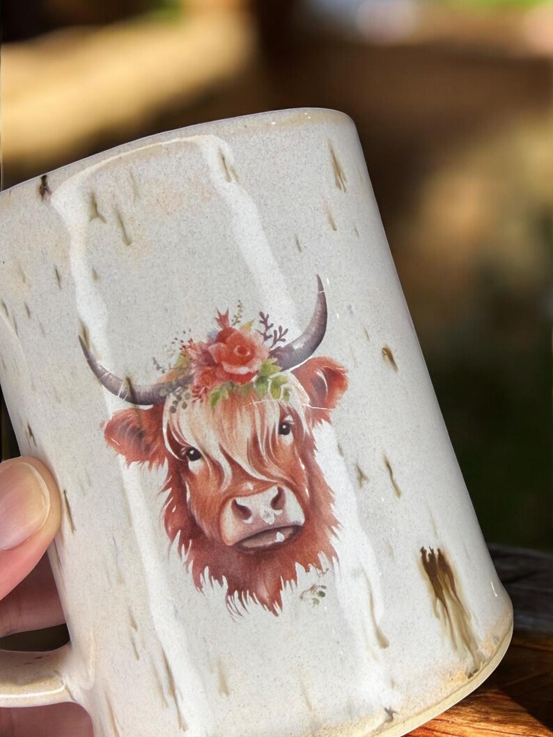 May include: Cream-colored ceramic mug featuring a Highland cow illustration. The cow has reddish-brown fur, a floral crown, and curved horns. The mug has a speckled design with brown drips.