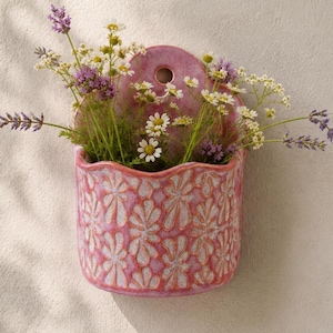 May include: A pink ceramic wall pocket planter featuring a repeating embossed floral pattern and a circular hole at the top for mounting. The planter is filled with a bouquet of white daisies, purple lavender, and small yellow wildflowers.