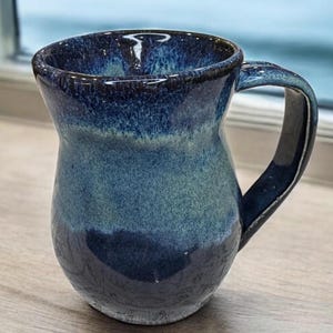 Könnte beinhalten: Eine handgefertigte Keramiktasse mit blauer Glasur. Die Tasse hat einen abgerundeten Körper und einen großen Henkel. Die Farbe geht von Dunkelblau am Boden zu helleren Blautönen am oberen Rand über. Die Tasse ist etwa 10 cm hoch.