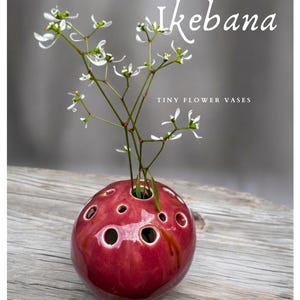May include: A small, red, ceramic vase with multiple holes for holding flowers. The vase is shaped like an egg and has a glossy finish. The vase is sitting on a wooden surface with a small bouquet of white flowers in it. The text "Ikebana Tiny Flower Vases handmade - for dried or fresh flowers" is visible below the vase.
