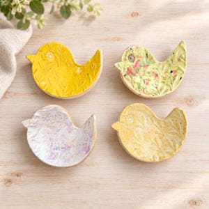 May include: Four bird-shaped ceramic trinket dishes. One is bright yellow with embossed floral patterns, one is pale yellow with pink and green floral details, one is white with purple speckles, and one is light yellow with embossed leaf patterns.