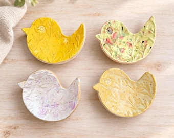 May include: Four bird-shaped ceramic trinket dishes. One is bright yellow with embossed floral patterns, one is pale yellow with pink and green floral details, one is white with purple speckles, and one is light yellow with embossed leaf patterns.