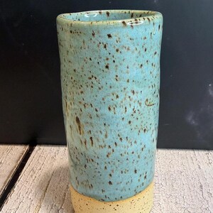 May include: A cylindrical ceramic tumbler featuring a glossy light blue glaze with dark brown speckles on the upper portion and a matte, speckled cream-coloured base. The vessel has a straight profile and a smooth, rounded rim at the top opening.