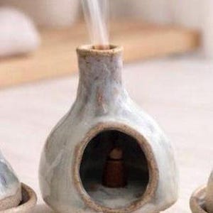 May include: A light blue and white ceramic incense burner with a rounded body, a central arched opening, and a narrow chimney top. A small brown incense cone sits inside the chamber, and a thin stream of white smoke rises from the top opening.