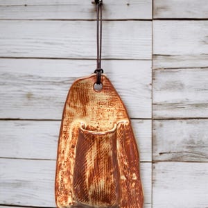 Könnte beinhalten: Eine handgefertigte Wandtasche aus Keramik mit rustikaler, strukturierter Oberfläche. Die Tasche ist in warmem, erdigem Braun mit helleren beigen Streifen gehalten und hat eine kleine Öffnung zur Aufbewahrung. Sie hängt an einer braunen Lederschnur.