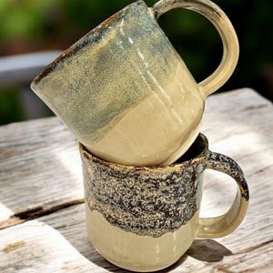 May include: Two handmade ceramic mugs stacked on a wooden surface. The top mug has a light blue and tan glaze, while the bottom mug features a speckled dark brown and tan glaze. Both mugs have rounded handles and a rustic aesthetic.