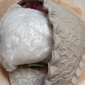 May include: A light gray ceramic bowl with a ruffled edge and leaf pattern. The bowl is partially filled with a white plastic bag. The bowl is unglazed and has a matte finish.