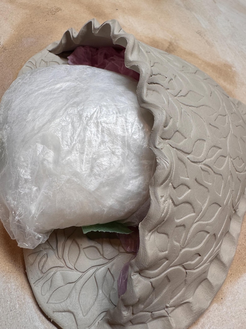 May include: A light gray ceramic bowl with a ruffled edge and leaf pattern. The bowl is partially filled with a white plastic bag. The bowl is unglazed and has a matte finish.