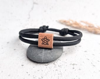 Könnte beinhalten: Schwarzes Seilarmband mit einem quadratischen Holzperlen. Die Perle ist mit einem Schildkrötenmuster graviert. Das Armband liegt auf einem grauen Stein. Der Hintergrund ist weiß.