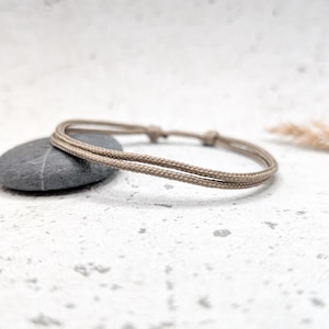 Könnte beinhalten: Ein schlichtes beigefarbenes Seil-Armband mit einem geknoteten Verschluss. Das Armband liegt auf einer weißen, strukturierten Oberfläche mit einem grauen Stein.