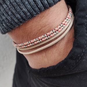 Può includere: Primo piano di tre braccialetti su un polso. Il braccialetto superiore è un design a corda con accenti rossi, neri e bianchi. Gli altri due braccialetti sono beige tinta unita. La persona indossa un maglione grigio e pantaloni neri.