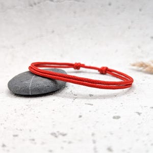 May include: A red string bracelet with a simple knot design. The bracelet is lying on a white surface with a gray stone.