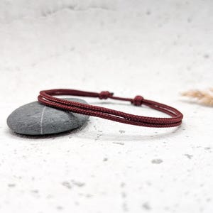 Könnte beinhalten: Ein schlichtes geflochtenes Armband aus burgunderrotem Kordel. Das Armband liegt auf einer weißen Oberfläche mit einem grauen Stein.