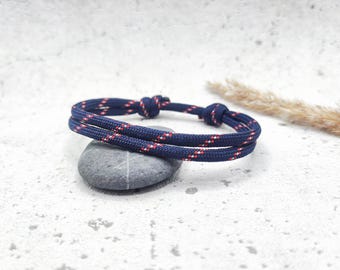 Può includere: Un braccialetto in corda blu navy e rosso con chiusura a nodo. Il braccialetto è su una superficie bianca con una pietra grigia.
