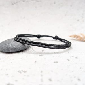 Könnte beinhalten: Ein schlichtes Armband aus geflochtenem schwarzem Kordel. Das Armband besteht aus zwei Kordelsträngen, die an den Enden miteinander verknotet sind.