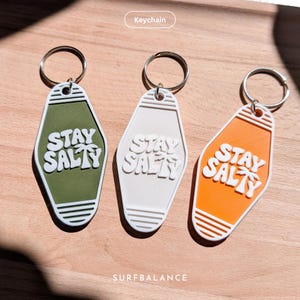 Puede incluir: Tres llaveros en verde, blanco y naranja, cada uno con un llavero plateado. Los llaveros tienen un diseño de llave de motel retro con las palabras "STAY SALTY" en letras blancas estilizadas. Un accesorio divertido.