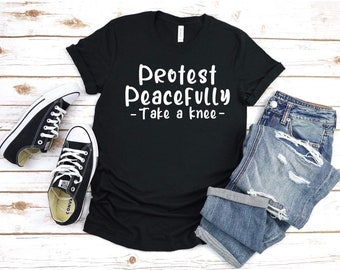 Pode incluir: Camiseta preta com texto branco que diz "Protest Peacefully - Take a knee -" A camiseta está dobrada e colocada sobre uma superfície de madeira branca com um par de tênis pretos e uma calça jeans azul.